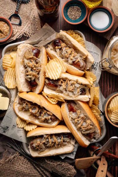 Tray of beer brats in buns topped with onions and grainy mustard, surrounded by ridged potato chips on a rustic table setting.