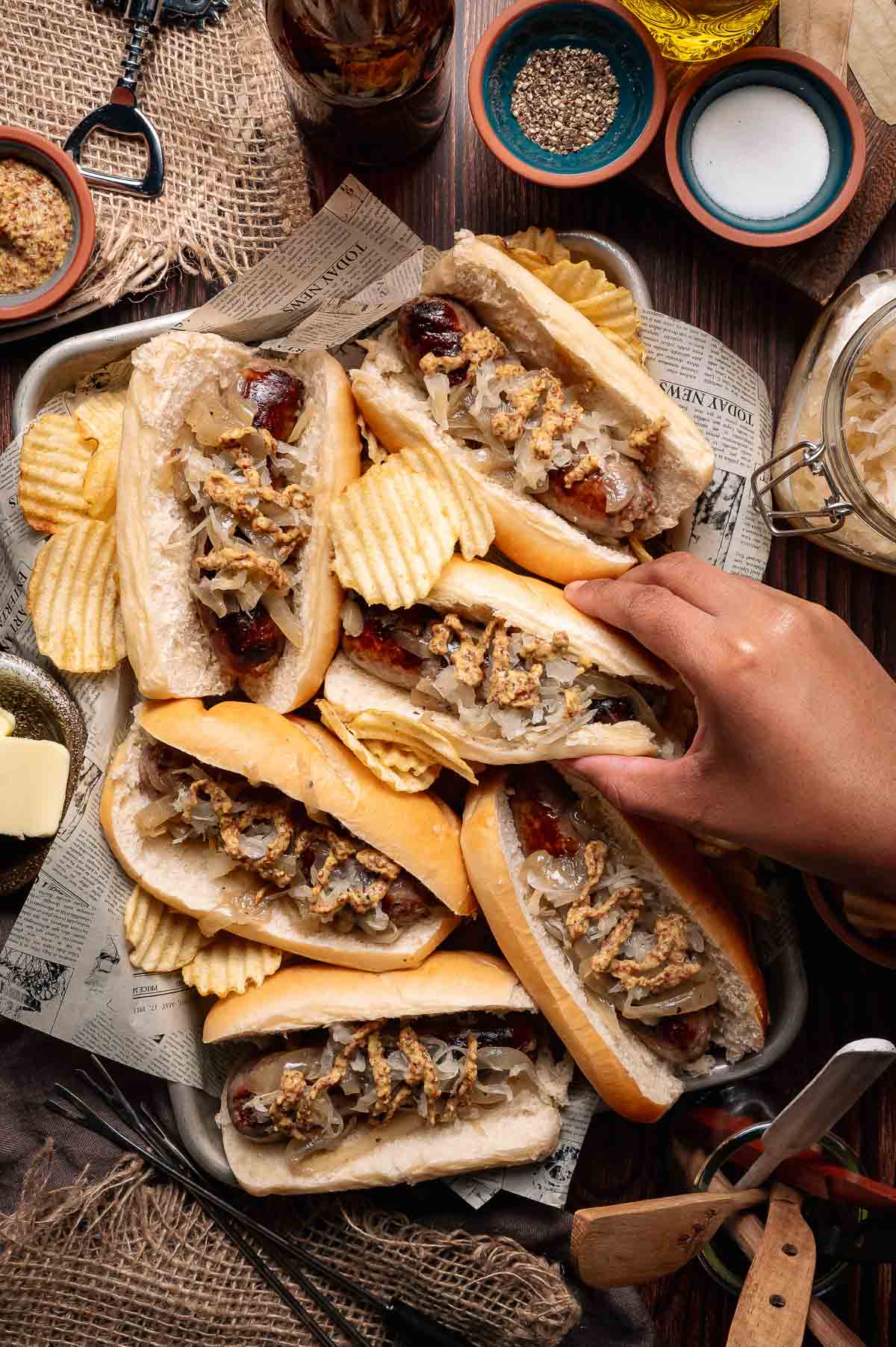 A hand reaches for a tray of beer brats in buns, topped with onions and mustard, served with potato chips on a newspaper-lined tray.