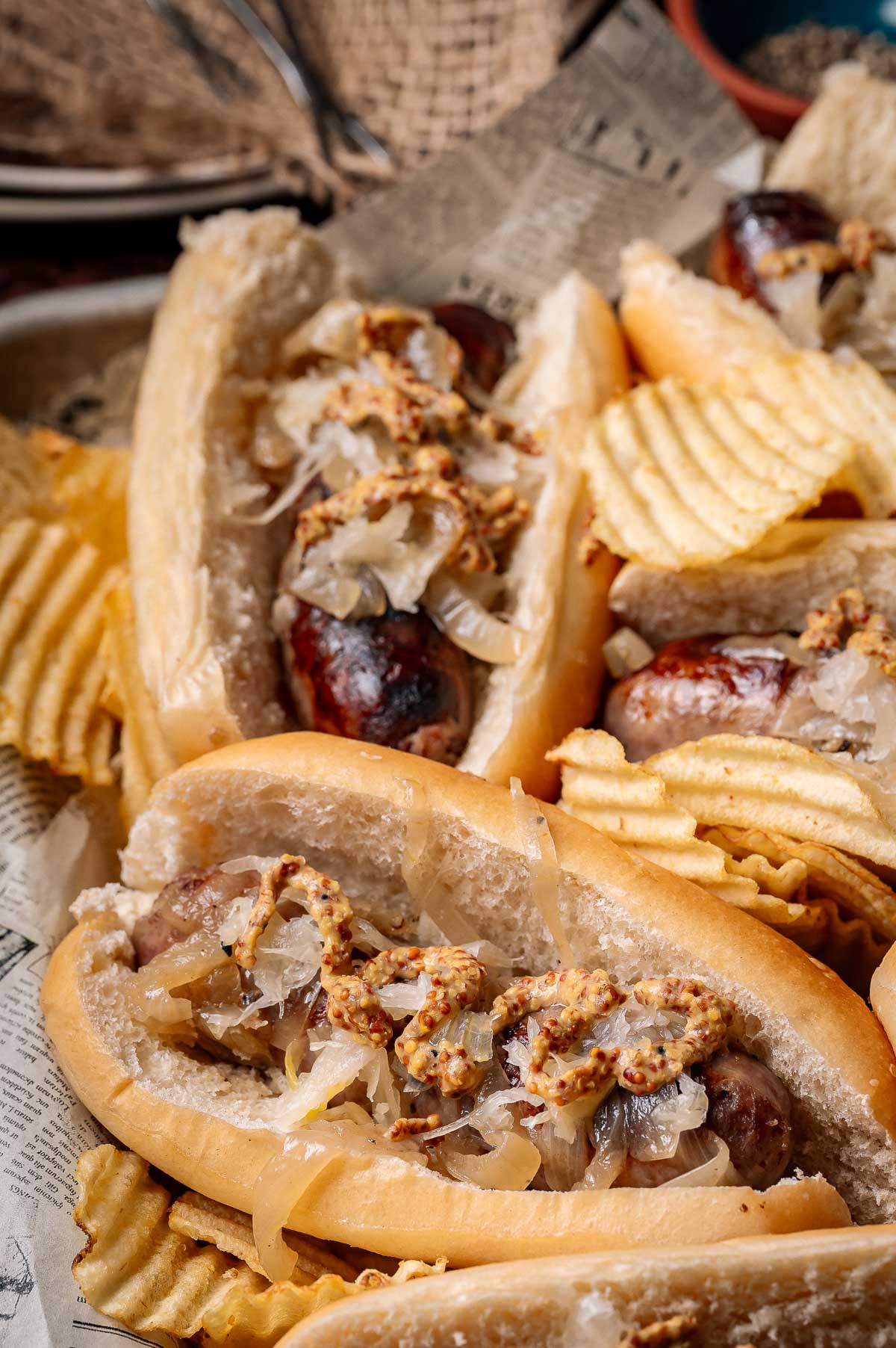 Three beer brats in buns topped with onions and grainy mustard, served with ridged potato chips on a sheet of newspaper.