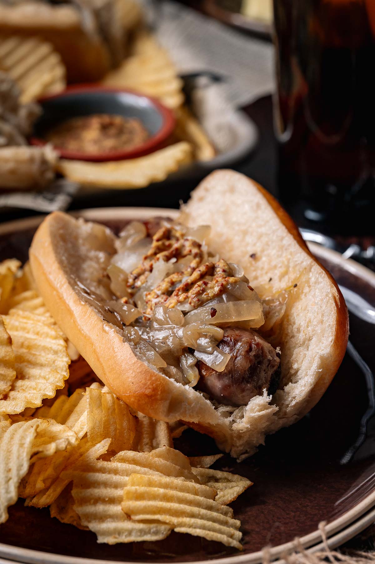 A beer brat in a bun topped with braised onions and mustard, served on a plate with ridged potato chips.