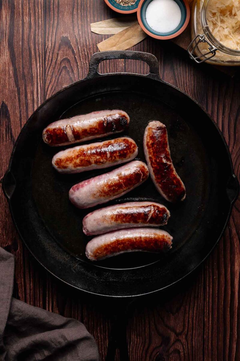 Six browned brats are cooking in a black cast iron skillet on a wooden surface, with sauerkraut, seasonings, and a cloth nearby.