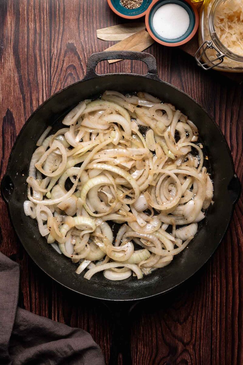 Sliced onions cooking in a black cast iron skillet on a wooden surface, with salt, pepper, and other ingredients nearby.