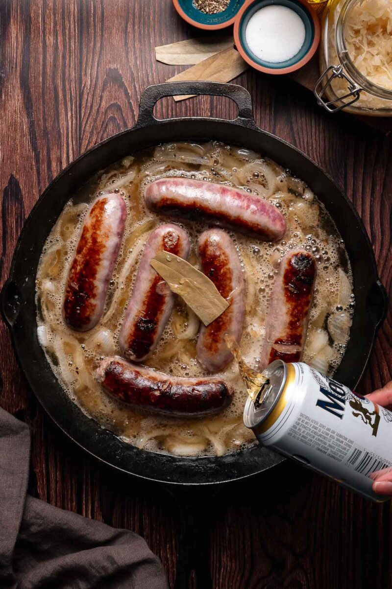Six brats are simmering in a cast iron skillet with onions and bay leaves while beer is being poured from a can over them on a wooden surface.