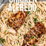 A skillet filled with fettuccine Alfredo pasta topped with sliced blackened chicken breasts and garnished with parsley. Text overlay: "How to Make Blackened Chicken Alfredo.