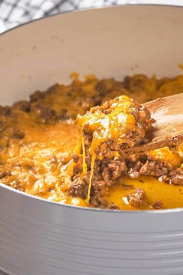 A skillet filled with cooked ground beef topped with melted cheddar cheese, being stirred with a wooden spoon.