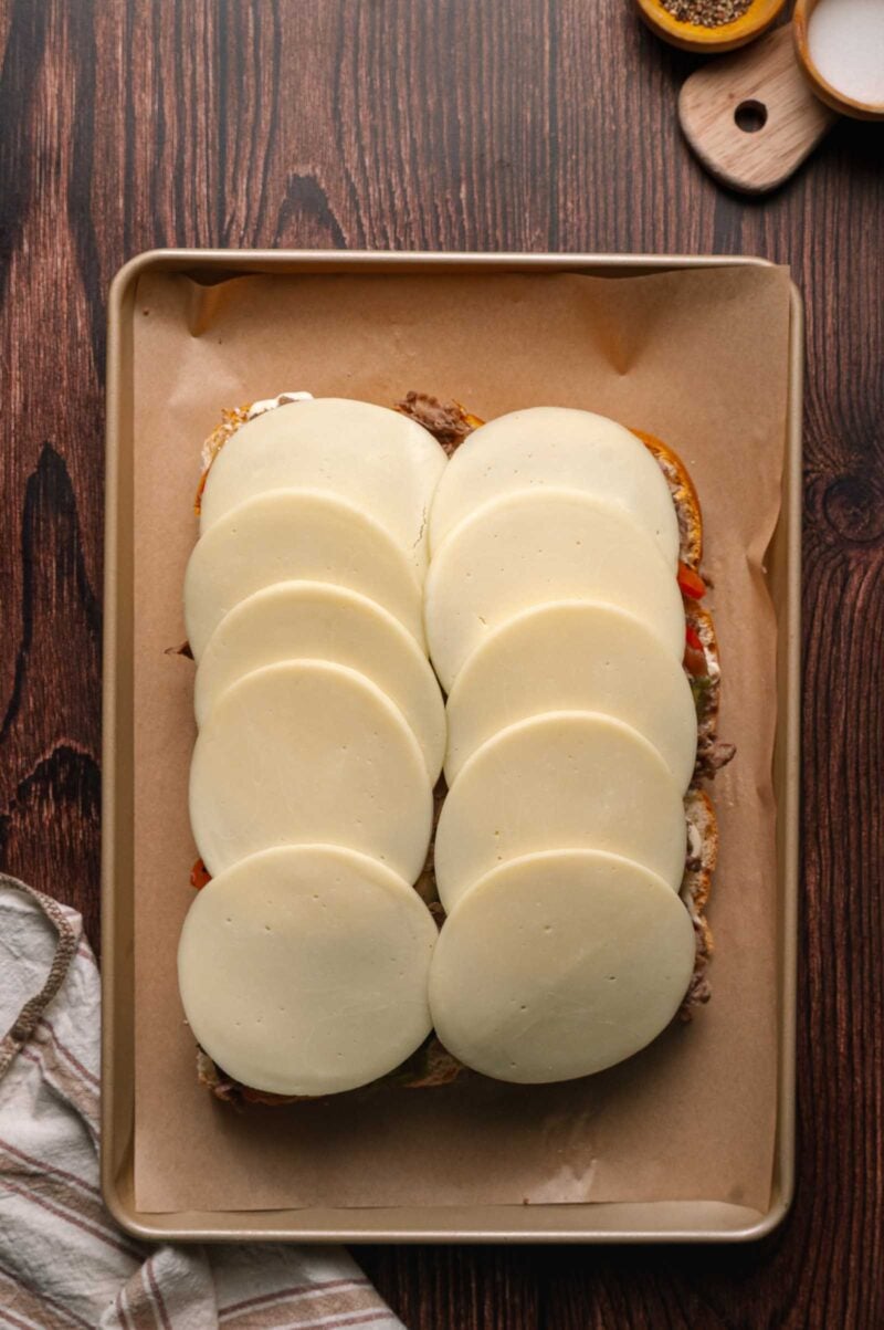 A baking sheet lined with parchment paper holds a cheesesteak sliders topped with overlapping slices of provolone cheese.