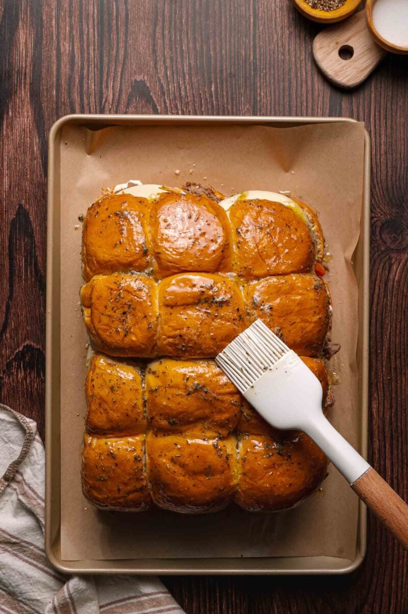 A tray of golden slider buns on parchment paper is being brushed with melted butter using a white silicone brush.