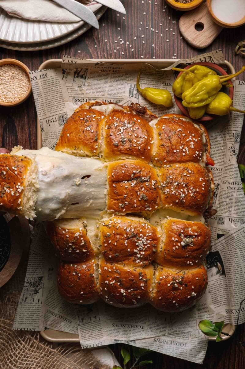 Golden pull-apart cheesesteak sliders topped with sesame seeds on a tray lined with newspaper, garnished with pickled peppers.
