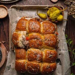A tray of pull-apart cheesesteak sliders topped with sesame seeds, filled with meat, served on newspaper with pepperoncini and seasonings on a wooden table.