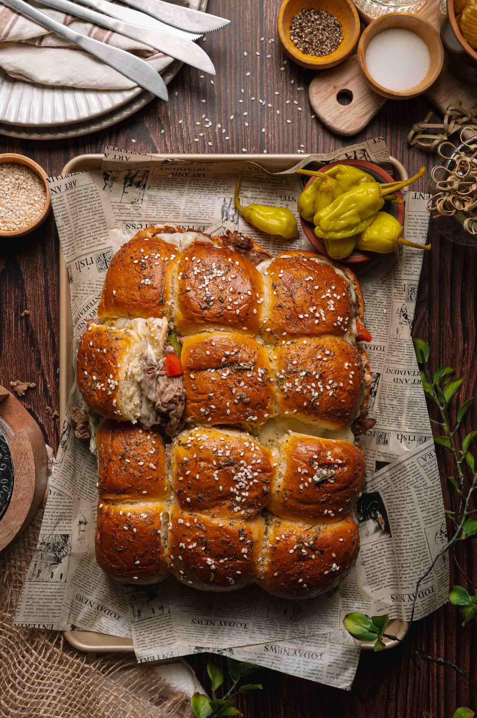 A tray of pull-apart cheesesteak sliders topped with sesame seeds, filled with meat, served on newspaper with pepperoncini and seasonings on a wooden table.