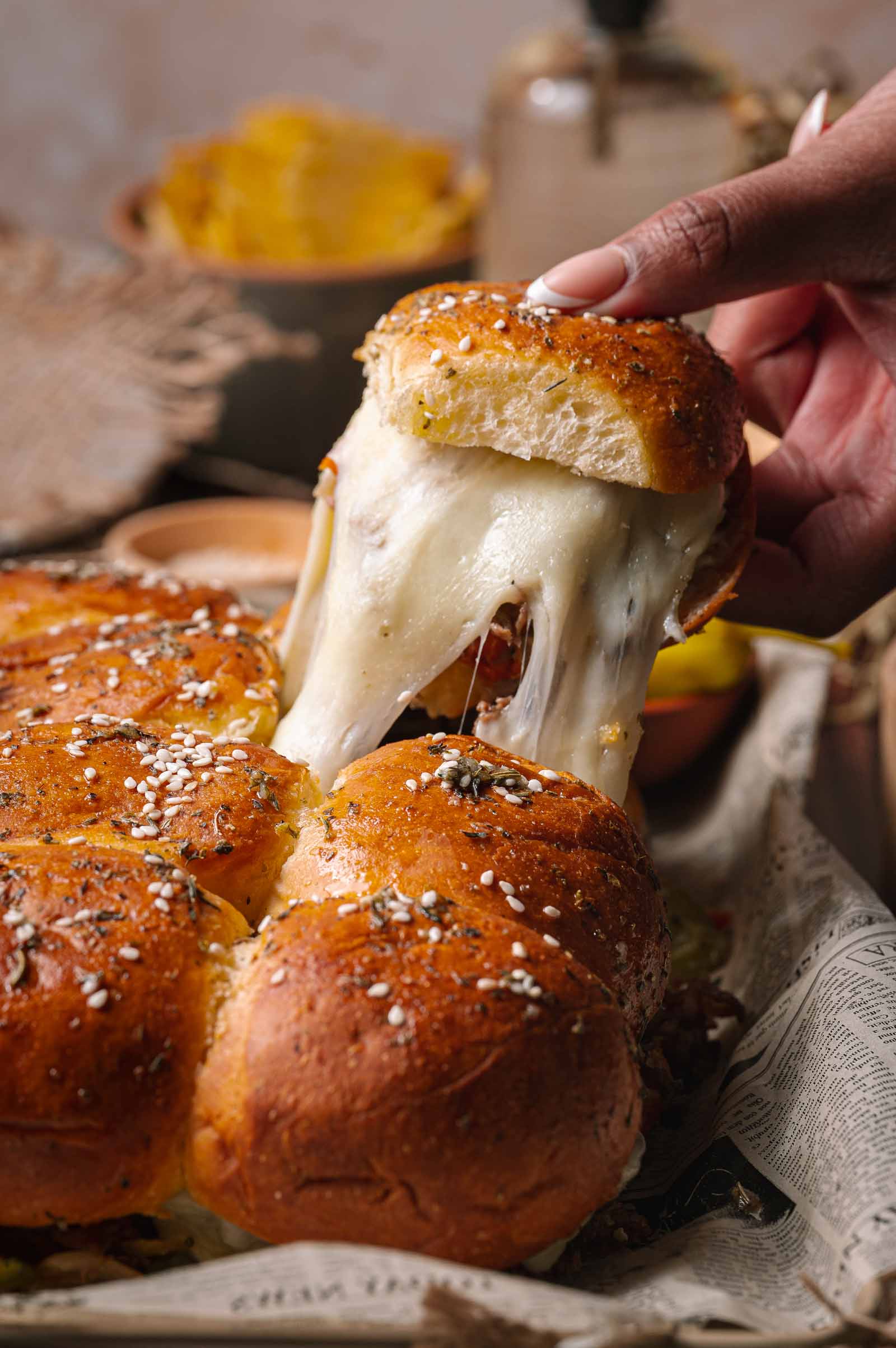 A hand pulls apart a golden, sesame-topped slider bun, stretching melted cheese from a tray of freshly baked cheesesteak sliders.