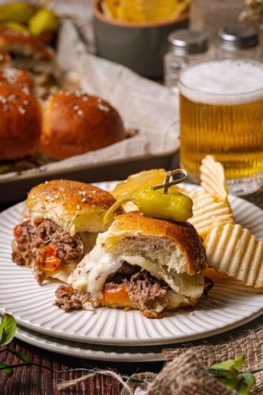 Two cheesy philly cheesesteak sliders on a plate with potato chips and a pickled pepper, with a glass of beer and more sliders in the background.