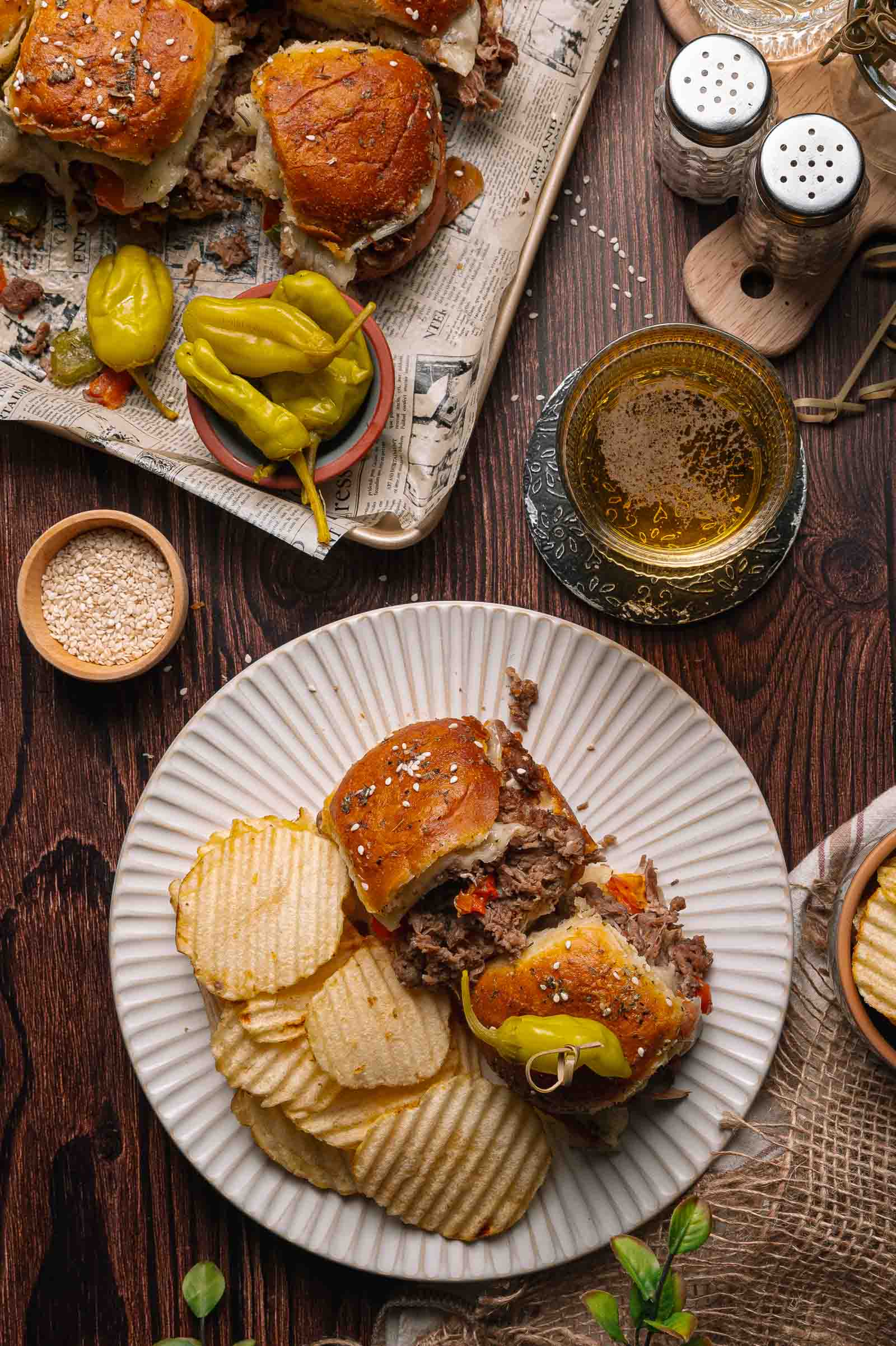 A plate with two cheesesteak sliders, ridged potato chips, and a pepperoncini; a tray with more sliders, pepperoncini, and seasonings is nearby on a wooden table.