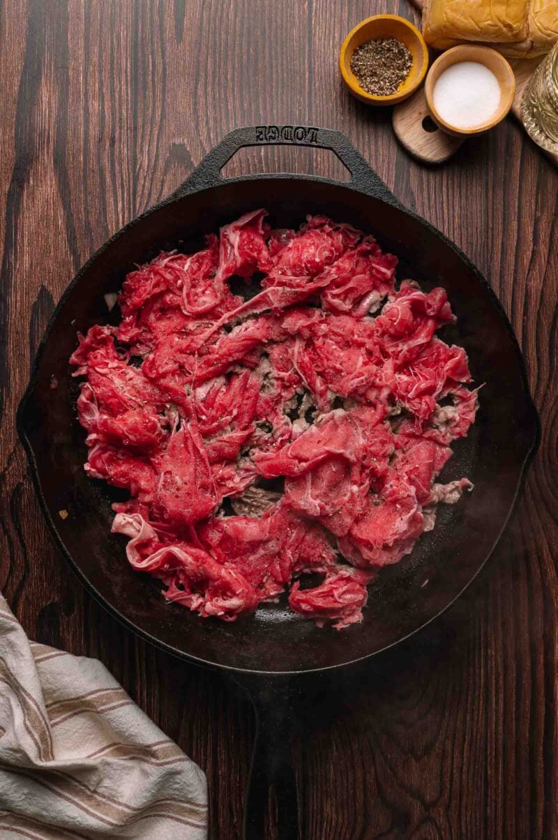 Thinly sliced raw ribeye steak cooking in a cast iron skillet on a wooden surface, with seasonings and oil nearby.