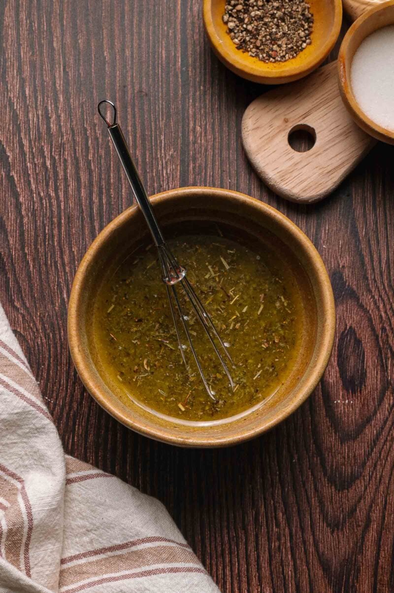 A ceramic bowl with seasoned butter and a whisk, placed on a wooden surface next to small bowls of salt and pepper and a striped cloth.