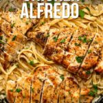 A skillet with sliced grilled chicken breast on top of fettuccine Alfredo pasta, garnished with chopped parsley. Text overlay reads "How to Make the Best Chicken Alfredo.
