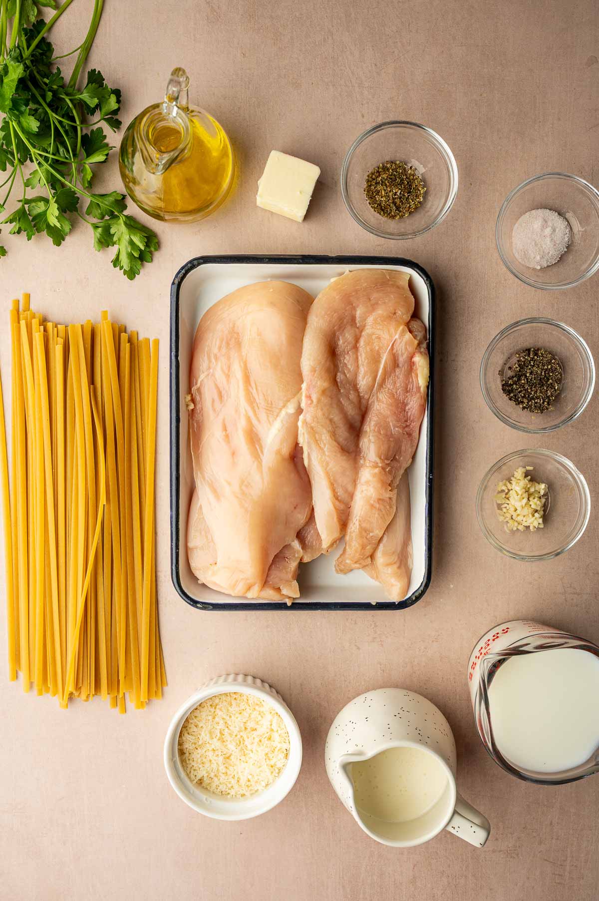 Raw chicken breasts, uncooked pasta, parsley, olive oil, butter, seasonings, minced garlic, grated cheese, milk, and cream arranged on a tan surface.