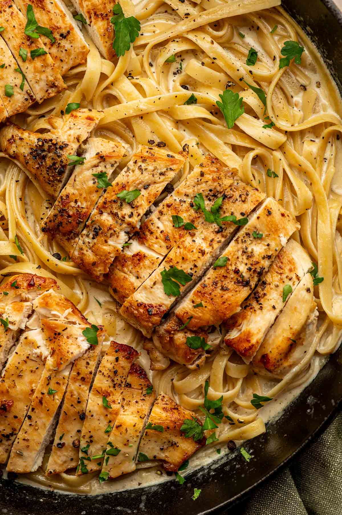 Sliced chicken breast served over creamy alfredo pasta, garnished with chopped parsley in a black pan.