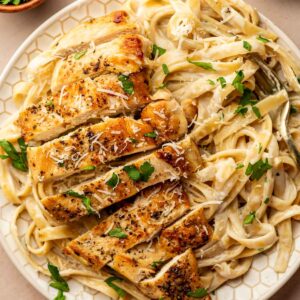Plate of fettuccine Alfredo topped with sliced chicken breast and garnished with chopped parsley, with bowls of grated cheese and parsley nearby.