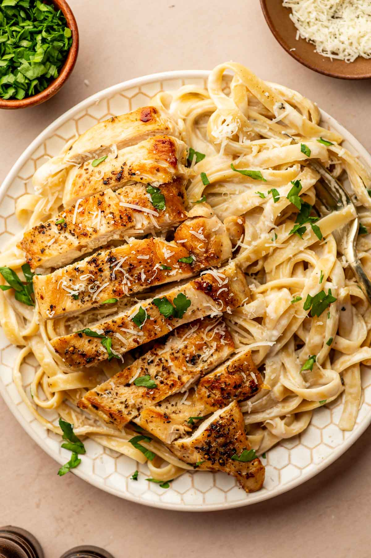 Plate of fettuccine Alfredo topped with sliced chicken breast and garnished with chopped parsley, with bowls of grated cheese and parsley nearby.