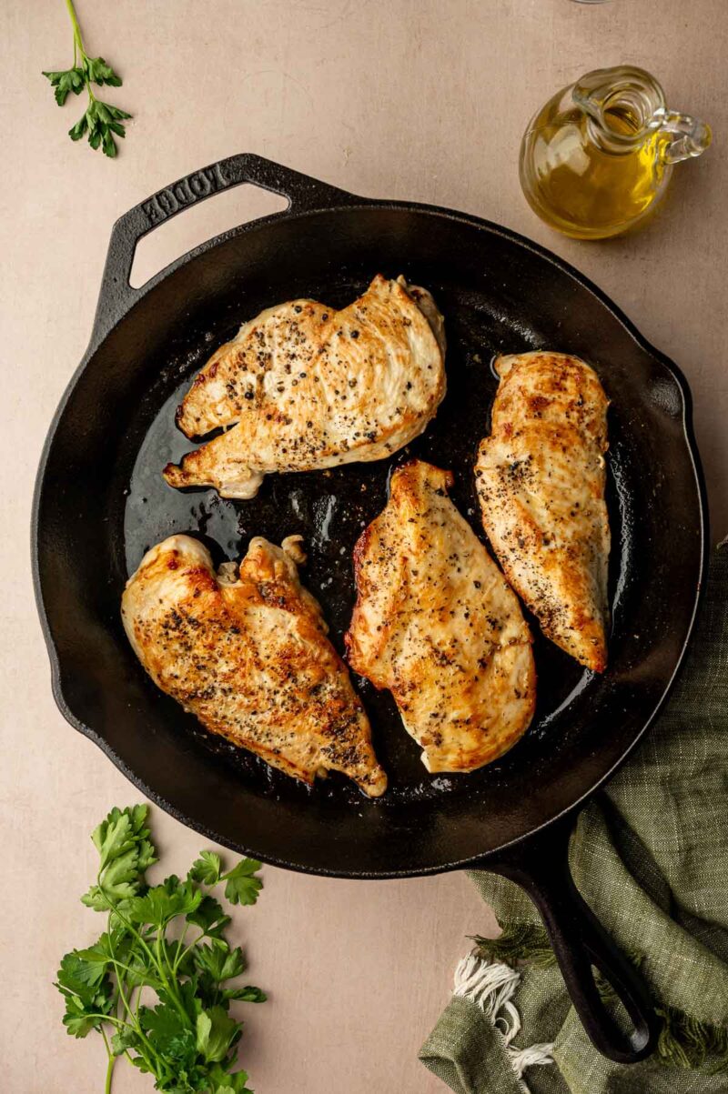 Four seasoned and cooked chicken breasts in a black cast iron skillet, with fresh parsley and a bottle of olive oil nearby.