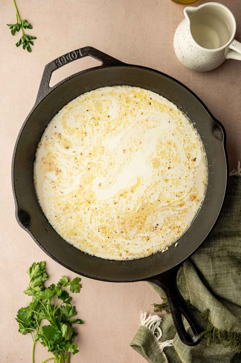 A cast iron skillet with a creamy white sauce and browned bits on the surface, surrounded by parsley, a green cloth, and a small white pitcher.