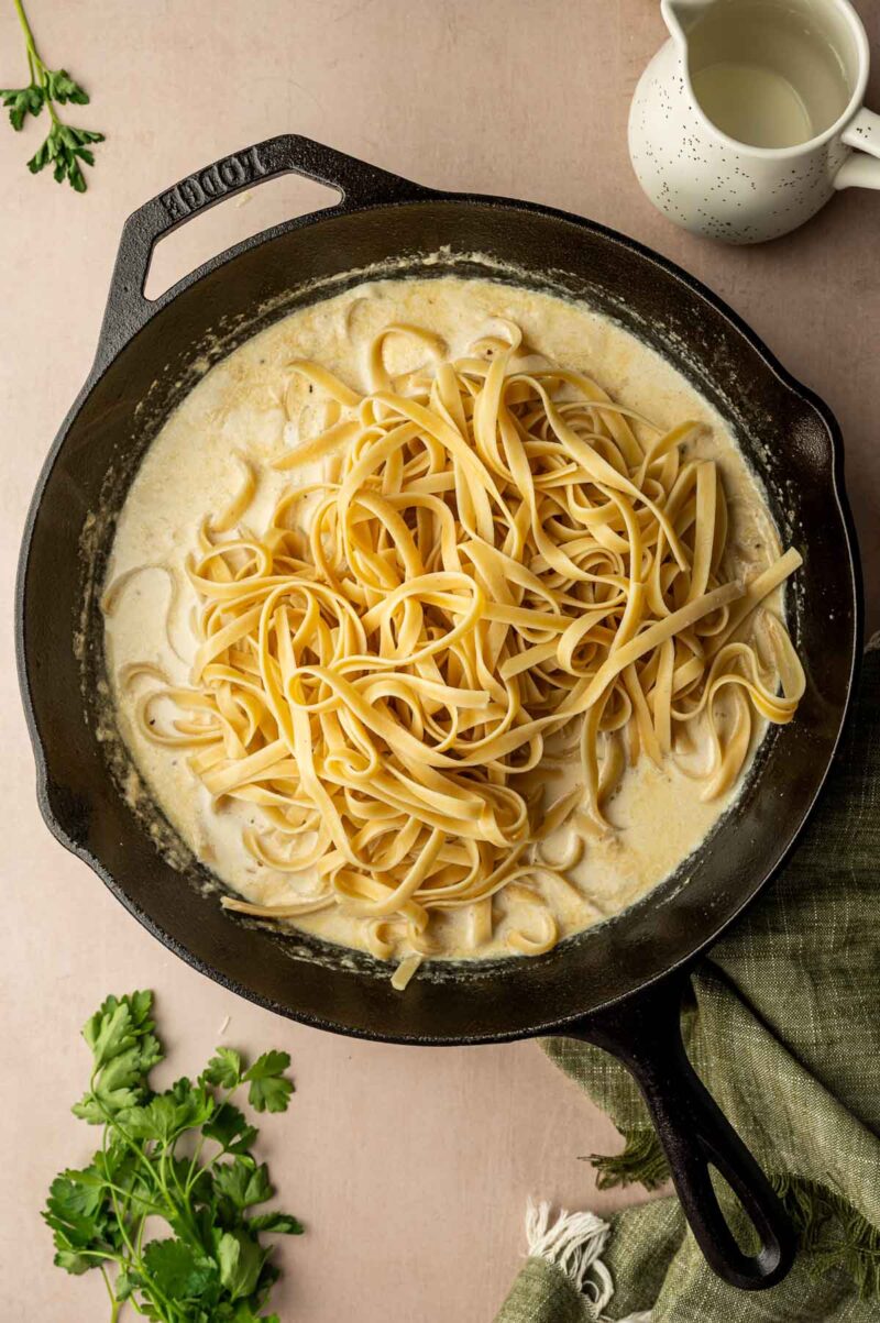 A cast iron skillet filled with creamy alfredo sauce and cooked fettuccine pasta, surrounded by fresh parsley and a small pitcher of cream.