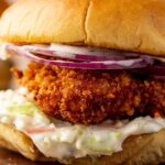 A close up of a golden crispy fried fish sandwich with coleslaw, sliced red onions, and mayonnaise on a soft bun to show off the ultra crispy panko coating.