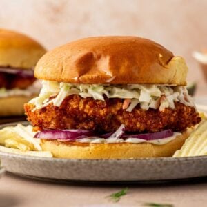 A fried fish sandwich with coleslaw and sliced red onions on a bun, served on a plate with potato chips.