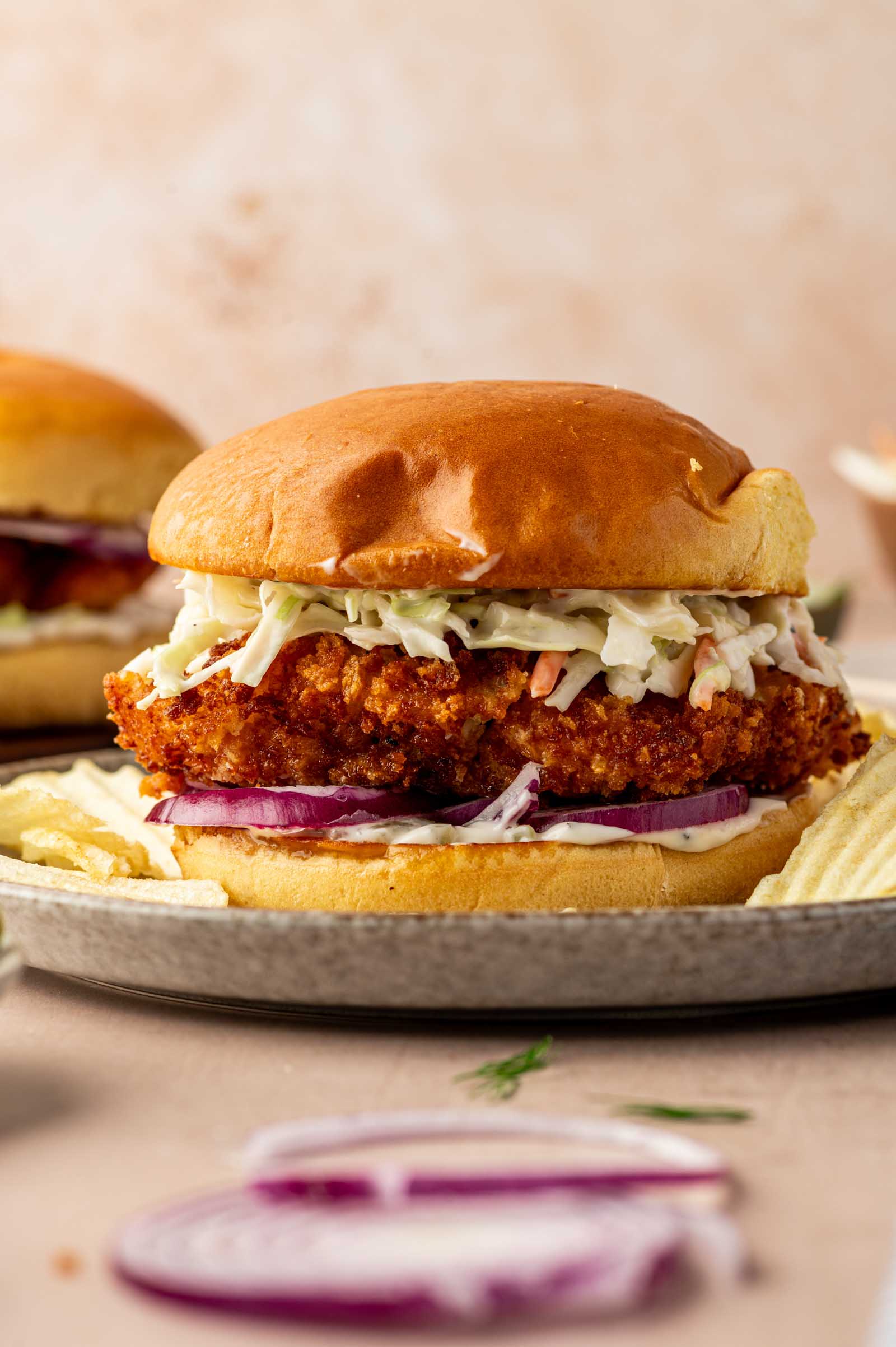 A fried fish sandwich with coleslaw and sliced red onions on a bun, served on a plate with potato chips.