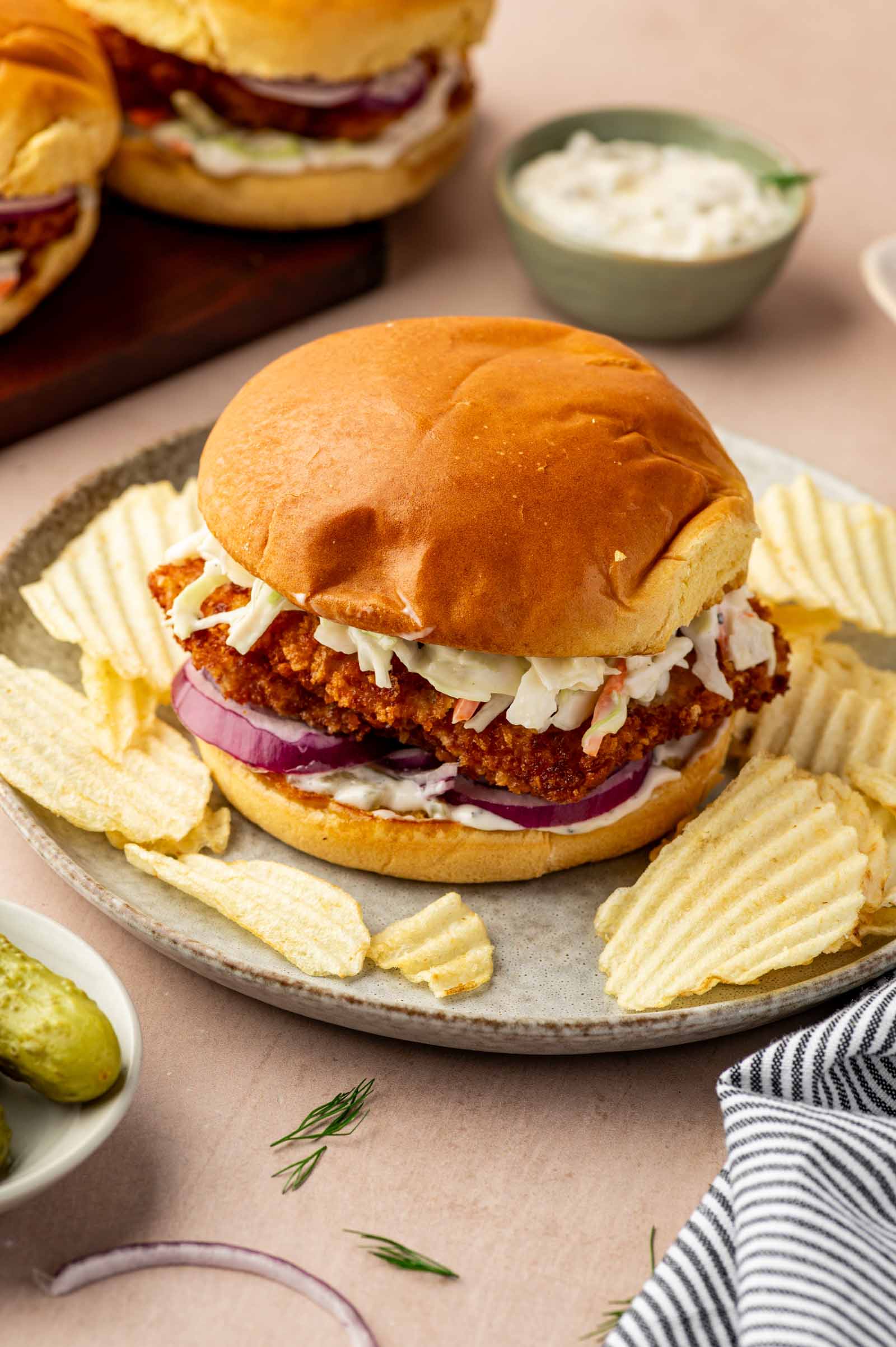 A fried fish sandwich with coleslaw and red onion on a bun, served with potato chips and pickles on the side.