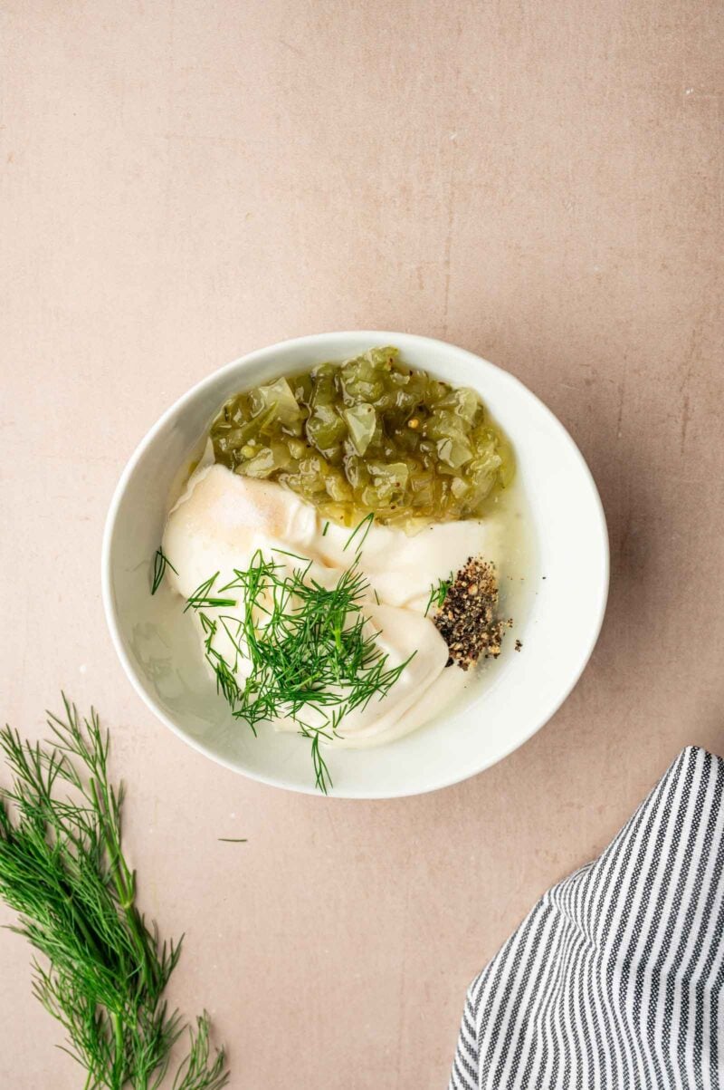 A white bowl containing mayonnaise, chopped pickles, black pepper, and fresh dill on a light surface, with a striped cloth and dill sprigs nearby.