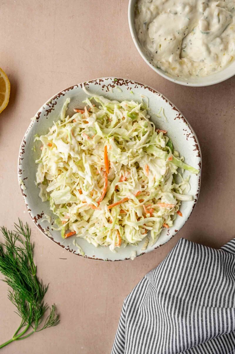 A bowl of creamy coleslaw sits on a light surface, next to a bowl of dressing, a striped cloth, dill sprigs, and a lemon wedge.