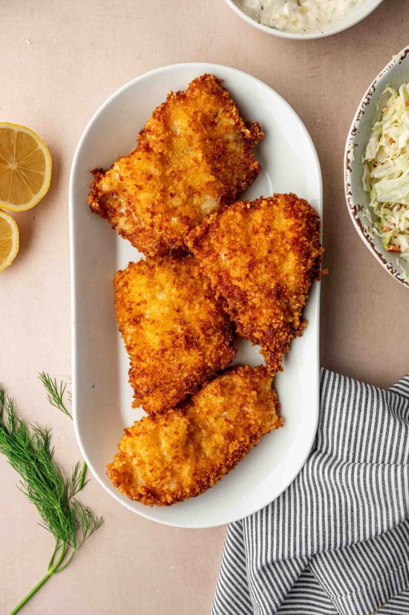 A white oval plate with four pieces of breaded and fried fish fillets, surrounded by sliced lemon, dill, coleslaw, and a striped napkin.