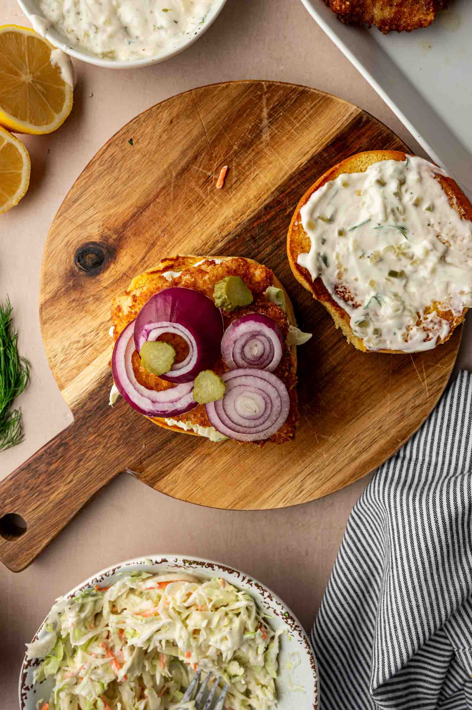 A fried fish sandwich with onions, pickles, and sauce on a wooden board, next to a bowl of coleslaw, lemon wedges, and a striped napkin.