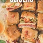 A batch of hot Italian sliders on sandwich rolls, filled with deli meats and topped with chopped vegetables, on a parchment-lined surface.