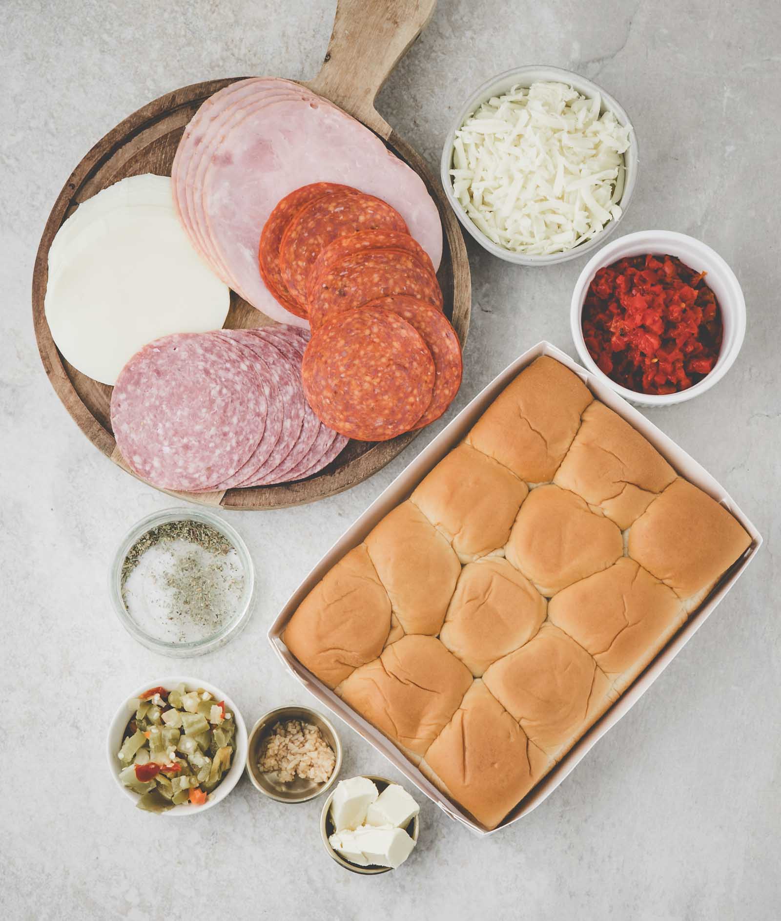 An assortment of deli meats and cheese on a wooden board, slider buns in a tray, and small bowls of shredded cheese, chopped peppers, seasonings, and condiments on a light surface.