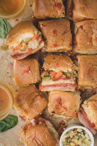 A top view of several hot italian sliders filled with cold cuts, cheese, giardiniera, and chopped vegetables on a parchment surface, with drinks and a small bowl of relish nearby.