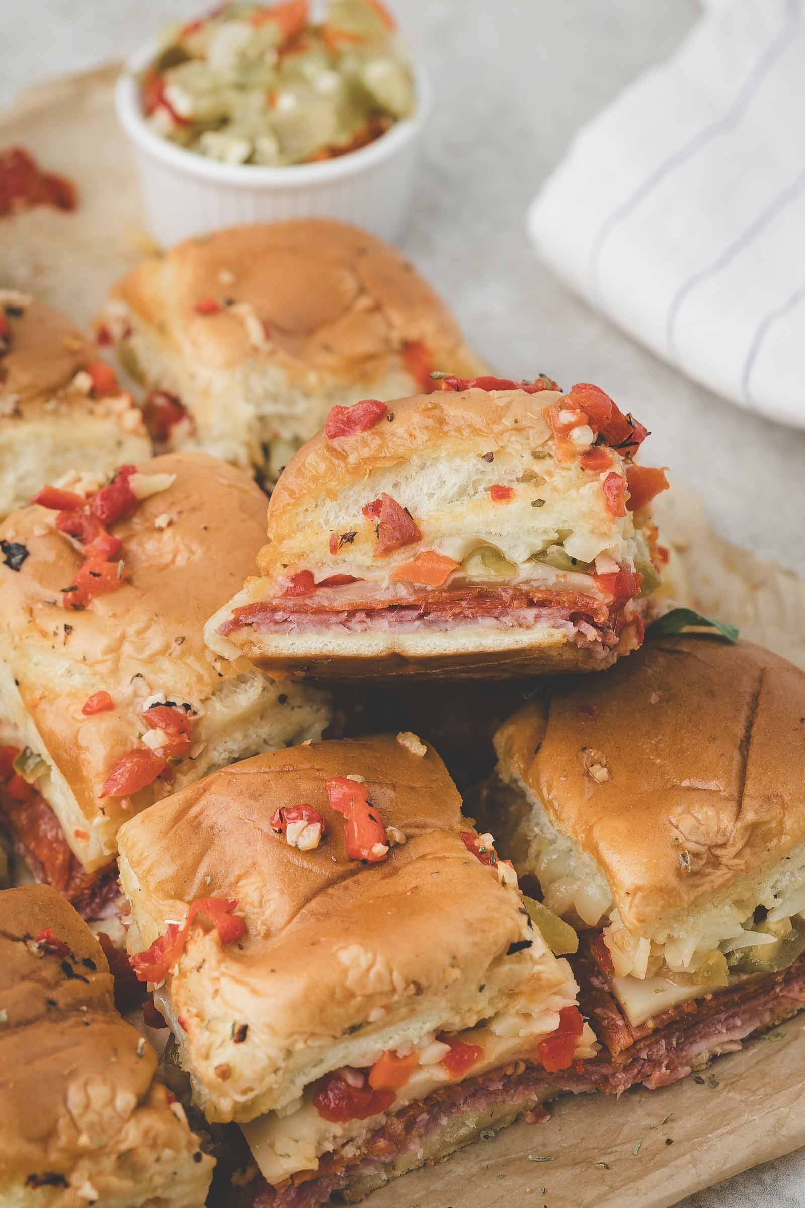 A stack of mini hot italian sliders with layers of deli meat, cheese, and chopped vegetables on soft buns, served on a wooden board with a small cup of relish in the background.
