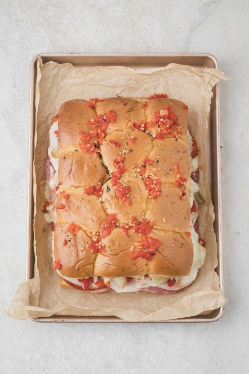 A baked slider loaf with melted cheese and diced red peppers on top, placed on parchment paper in a rectangular baking tray.