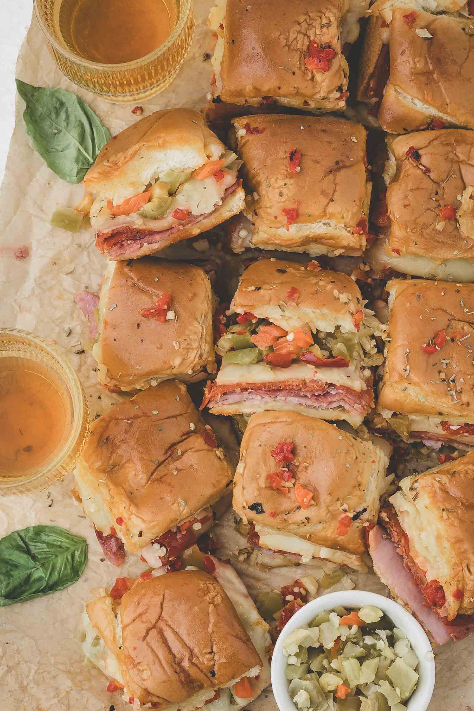 A dozen hot italian slider sandwiches with deli meats and pickles are arranged on parchment paper, accompanied by two drinks, basil leaves, and a small bowl of chopped vegetables.
