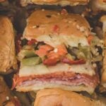 Close-up of a hot italian sub sandwich slider with layers of deli meat, cheese, and chopped pickled vegetables on a soft bun, surrounded by similar sandwiches.