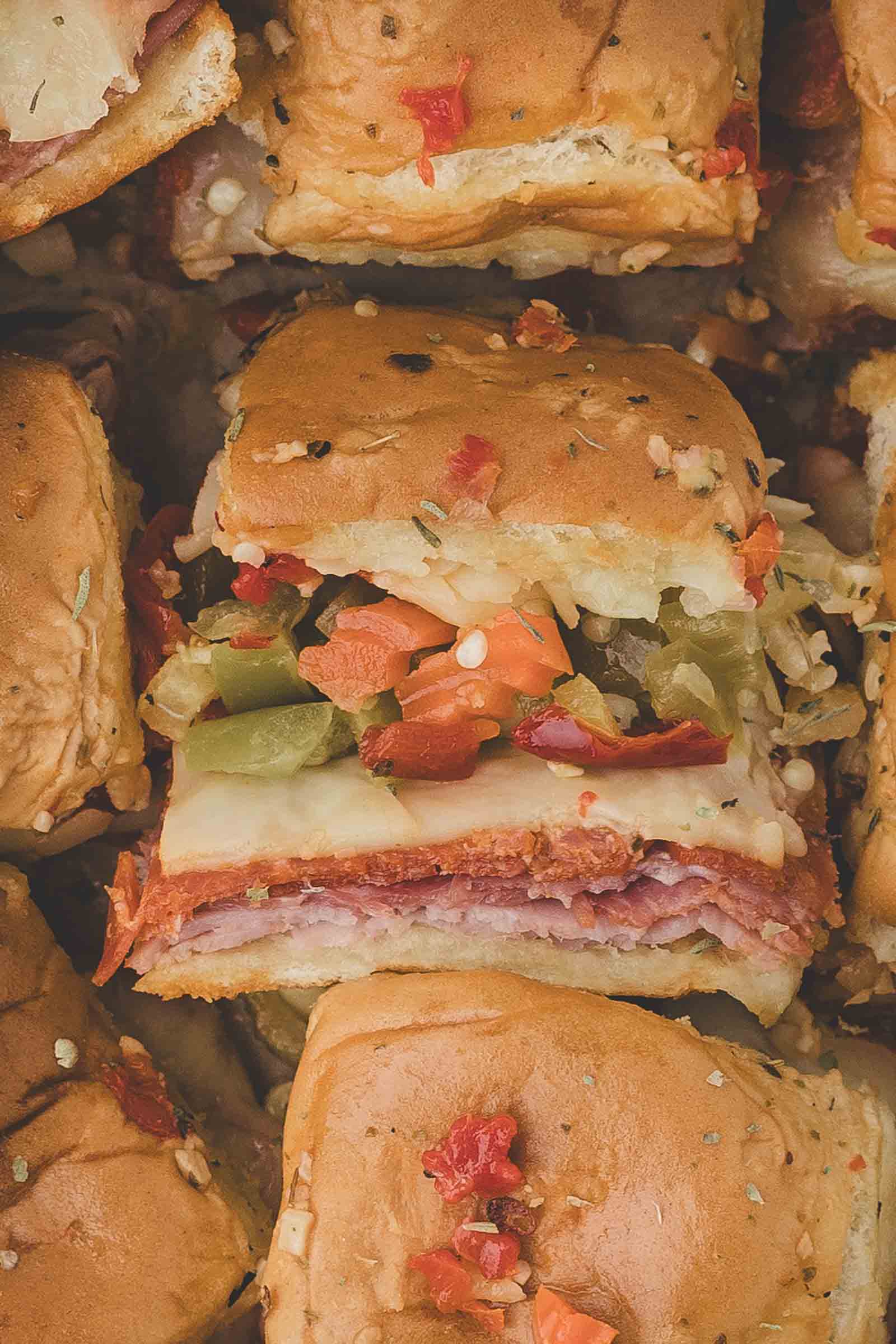 Close-up of a hot italian sub sandwich slider with layers of deli meat, cheese, and chopped pickled vegetables on a soft bun, surrounded by similar sandwiches.