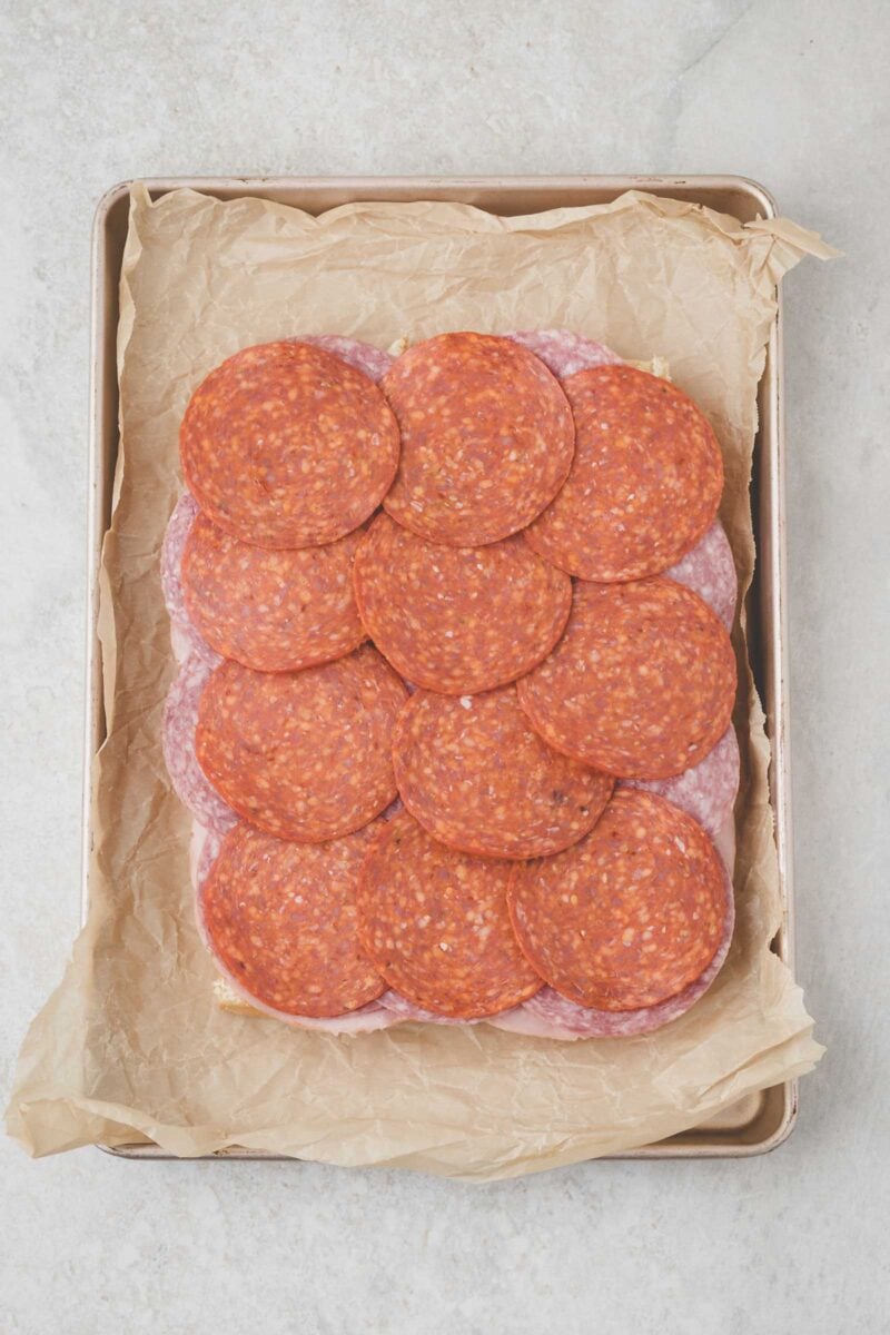 Rectangular baking tray lined with parchment paper, topped with overlapping slices of pepperoni and salami arranged on a layer of slier rolls.