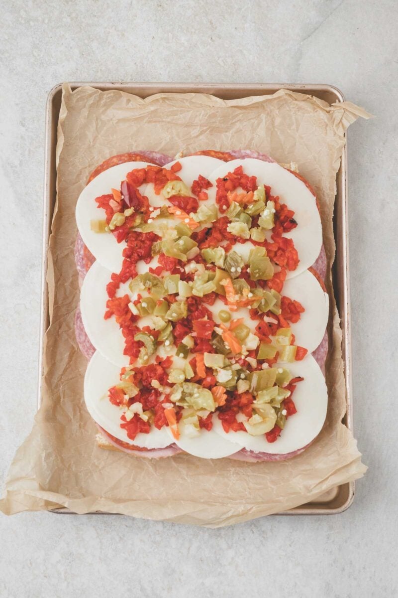 Deli meats and cheese layer Hawaiian rolls topped with sliced mozzarella, chopped Giardiniera on parchment-lined baking sheet.