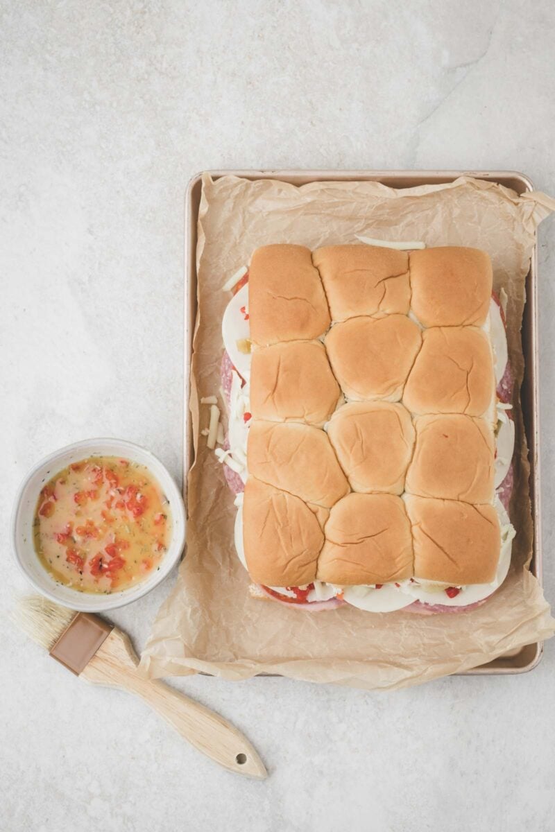 A tray of assembled slider sandwiches with a bowl of melted butter and seasonings, and a pastry brush on the side.