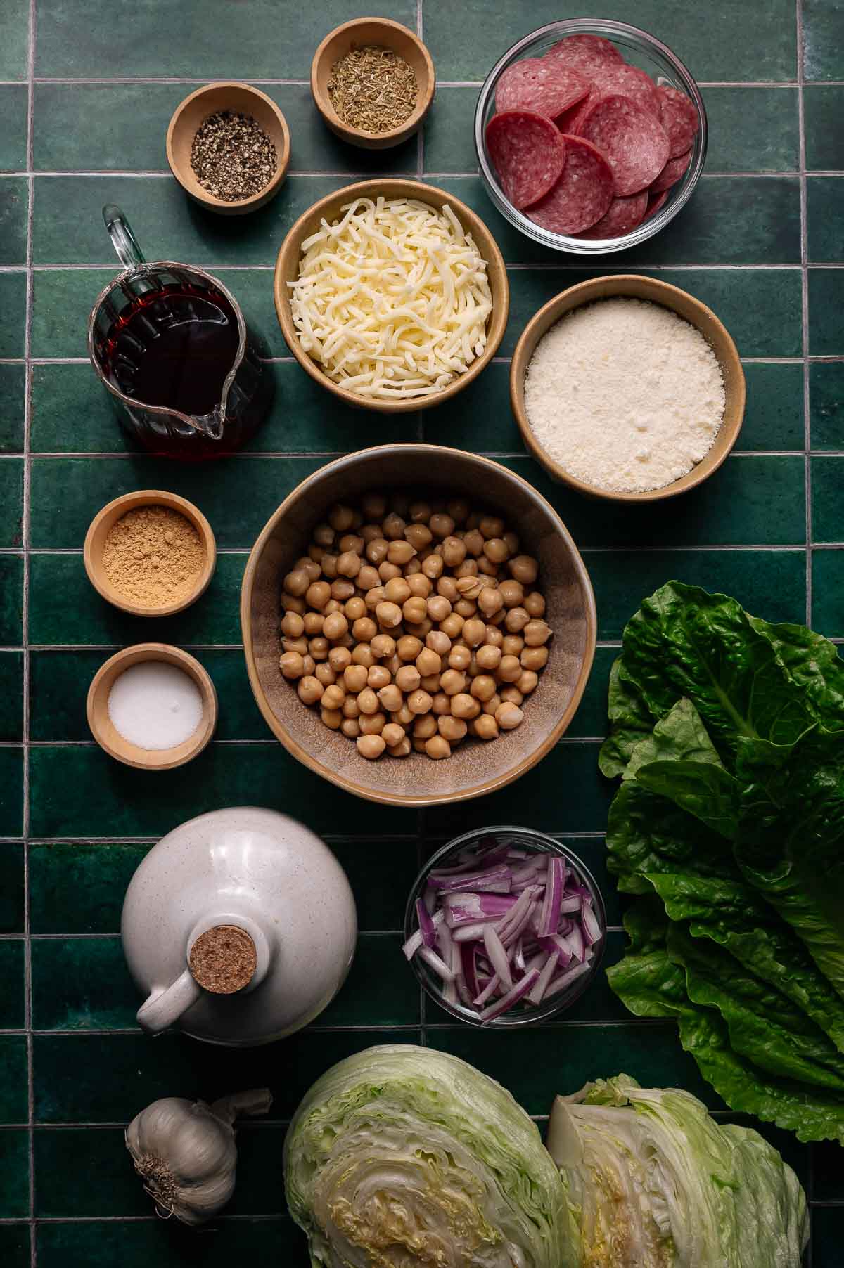 A flat lay of la scala salad ingredients including chickpeas, sliced salami, shredded cheese, grated cheese, red onion, lettuce, and various seasonings on a green tiled surface.