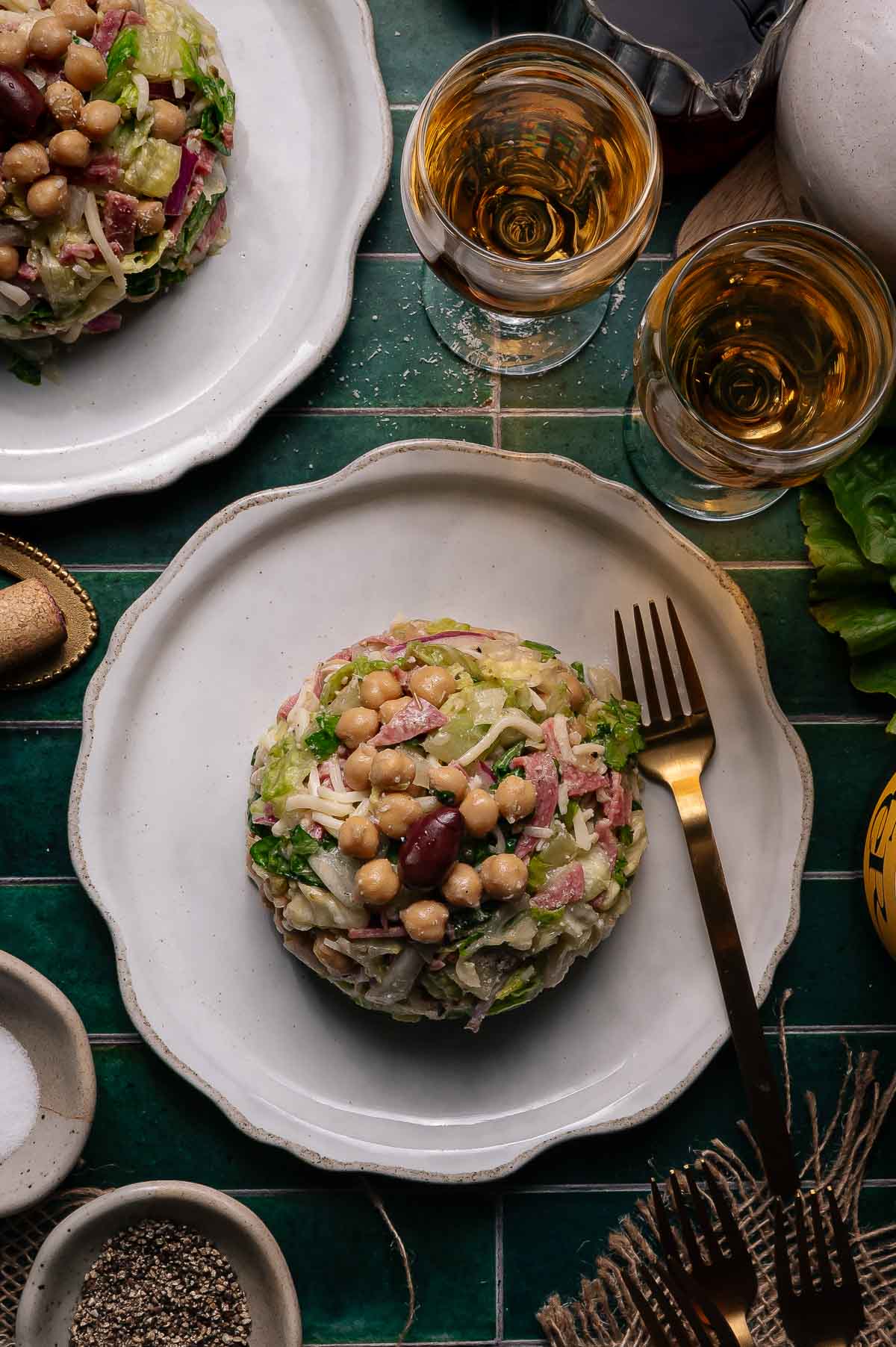 A composed La Scala chopped salad with chickpeas, the signature olive, red onions, and lettuce is served on a white plate with a fork; two glasses of amber-colored beverage are nearby on a green tile surface.