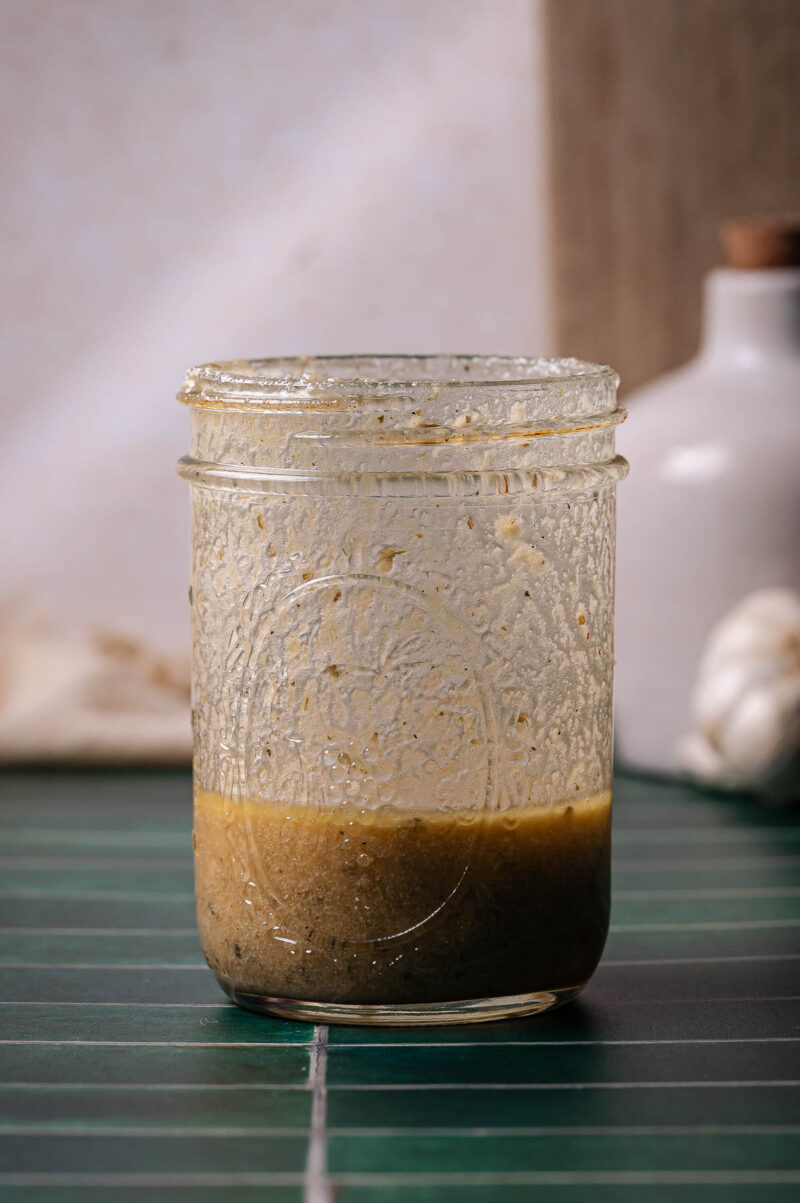A glass jar containing a vinaigrette sits on a green tiled surface with a white bottle and garlic in the blurred background.