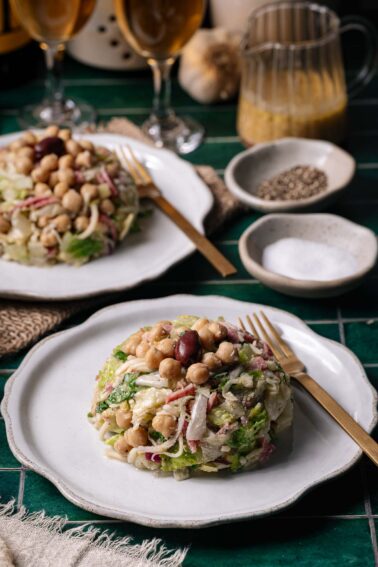 Two plates of La Scala salad with the signature olive on top, chickpeas, radishes, and greens on white dishes, set on a green tile table with cutlery, condiments, and drinks in the background.